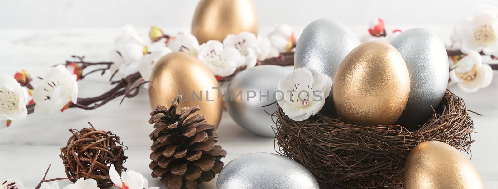 Close up of golden and silver Easter eggs in the nest with white plum flower on bright white wooden table background.
