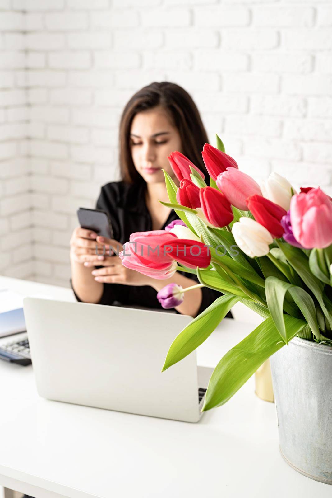 Small business concept. Young serious brunette business woman florist communicating on the phone, working at the office, bucket of tulips on the desk. Focus on flowers. Selective focus