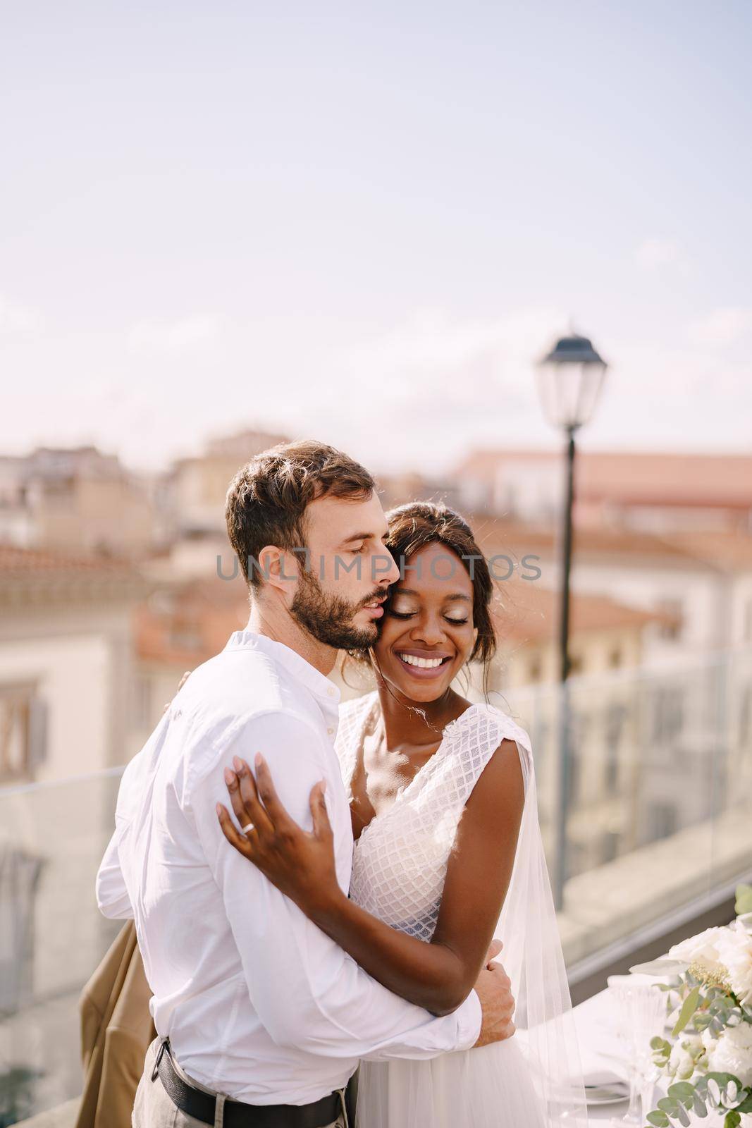 Multiethnic wedding couple. Destination fine-art wedding in Florence, Italy. Caucasian groom and African-American bride cuddling on a rooftop in sunset sunlight. by Nadtochiy