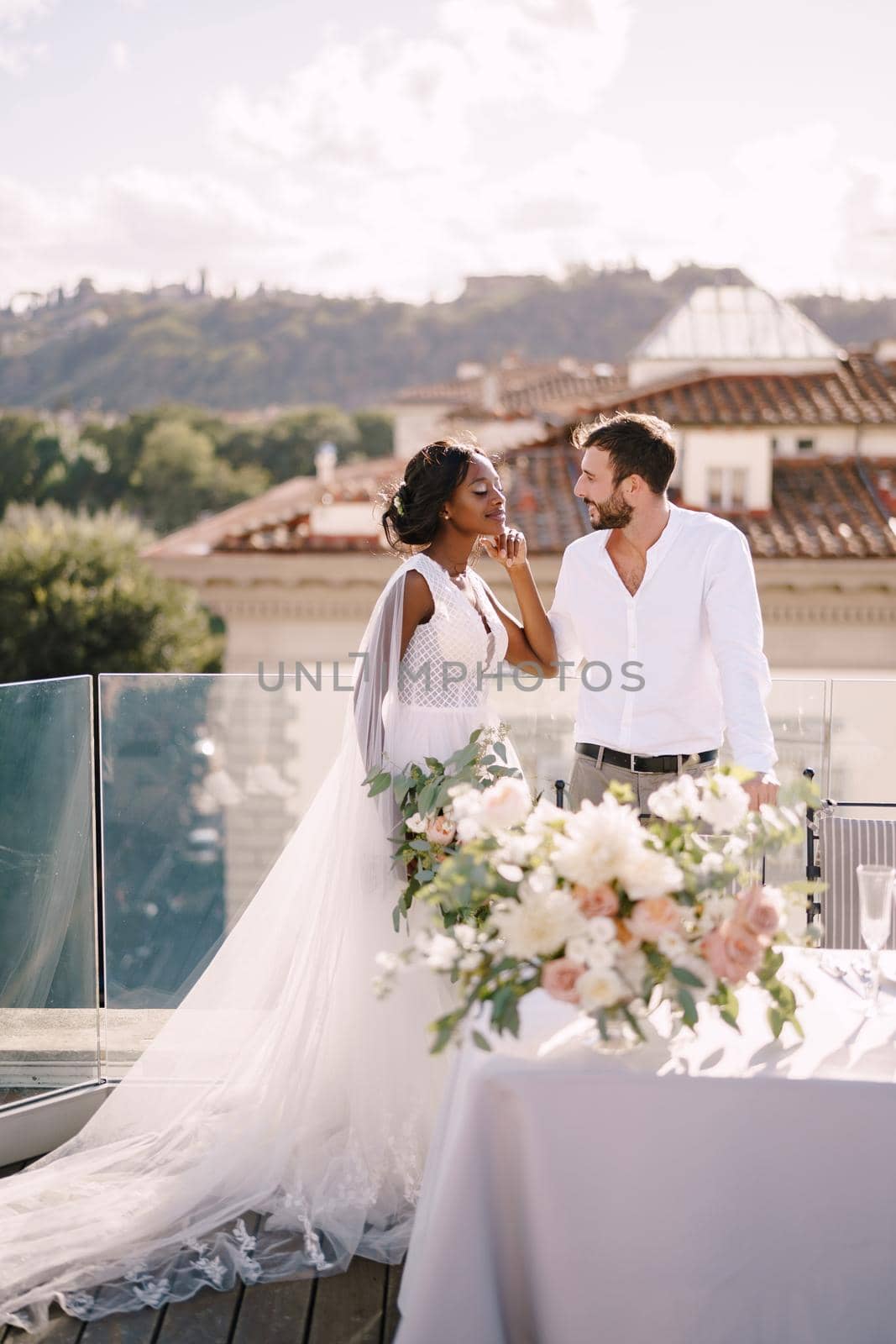 Destination fine-art wedding in Florence, Italy. Multiethnic wedding couple. African-American bride and Caucasian groom stand near the table for a wedding dinner. by Nadtochiy