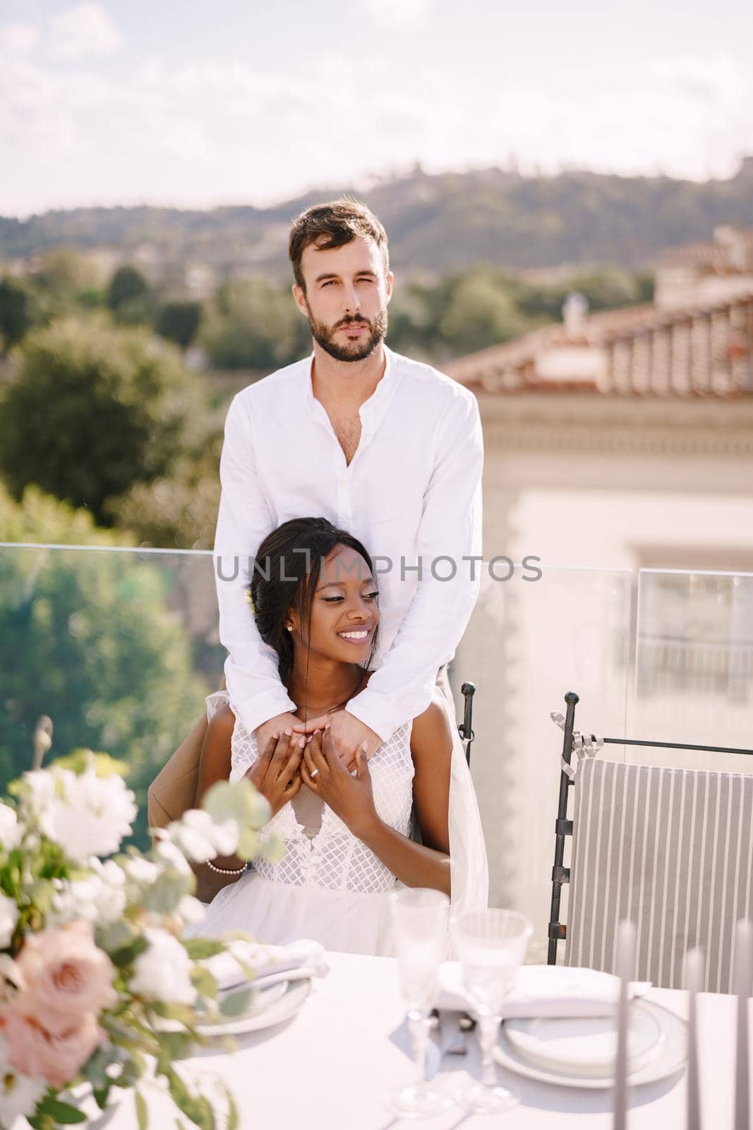 Interracial wedding couple. Destination fine-art wedding in Florence, Italy. African-American bride sits at wedding table, Caucasian groom hugs her shoulders.