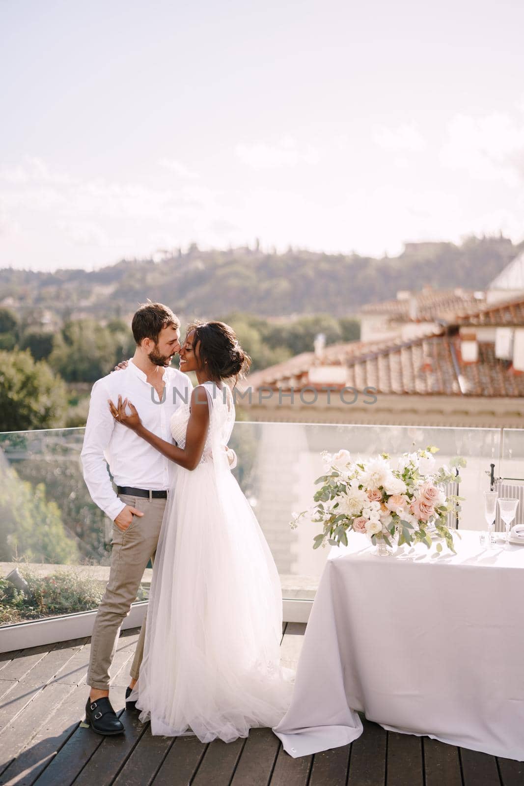 Interracial wedding couple. Destination fine-art wedding in Florence, Italy. African-American bride and Caucasian groom stand near the table for a wedding dinner. by Nadtochiy