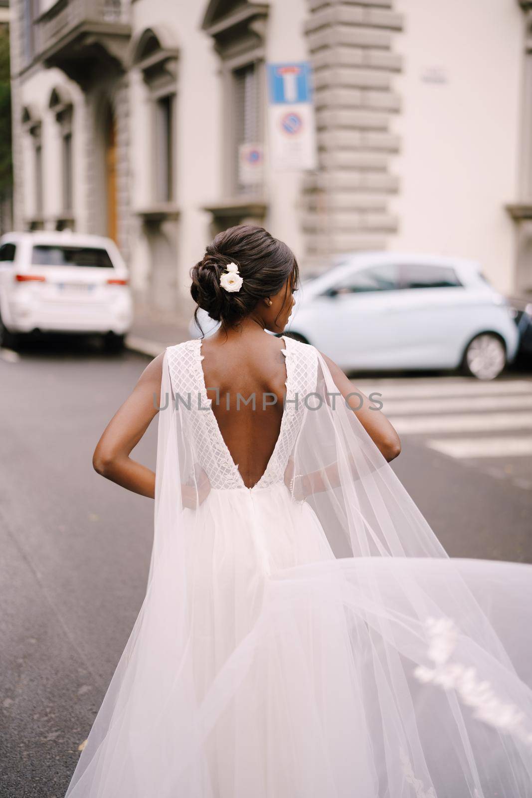 An African-American bride in a white dress, with a long veil, walks on the road on a city street. Wedding in Florence, Italy