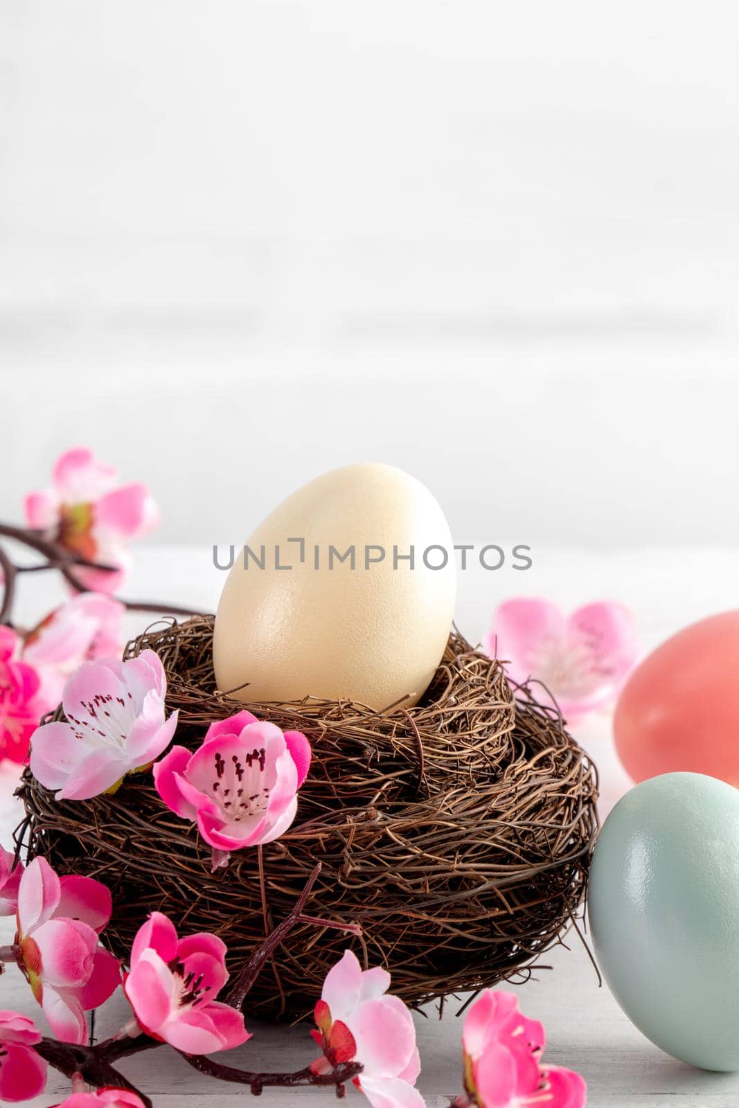Close up of colorful Easter eggs in the nest with pink plum flower on bright white wooden table background.