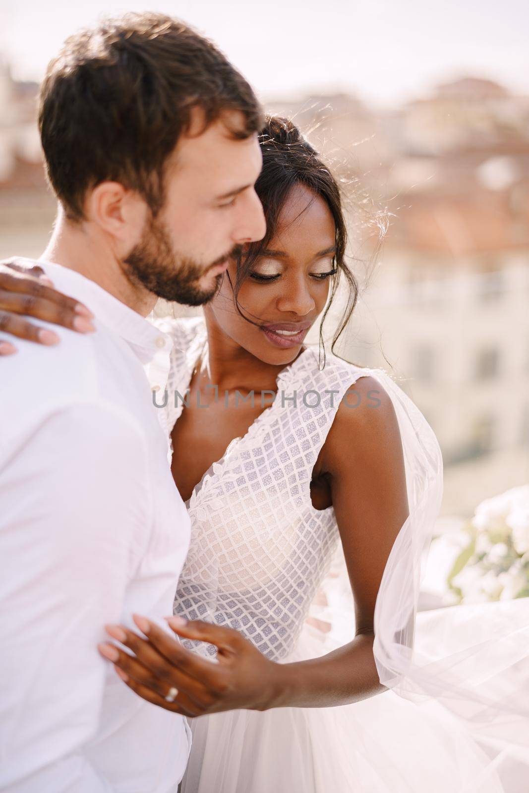 Destination fine-art wedding in Florence, Italy. Interracial wedding couple. Caucasian groom and African-American bride cuddling on a rooftop in sunset sunlight. by Nadtochiy