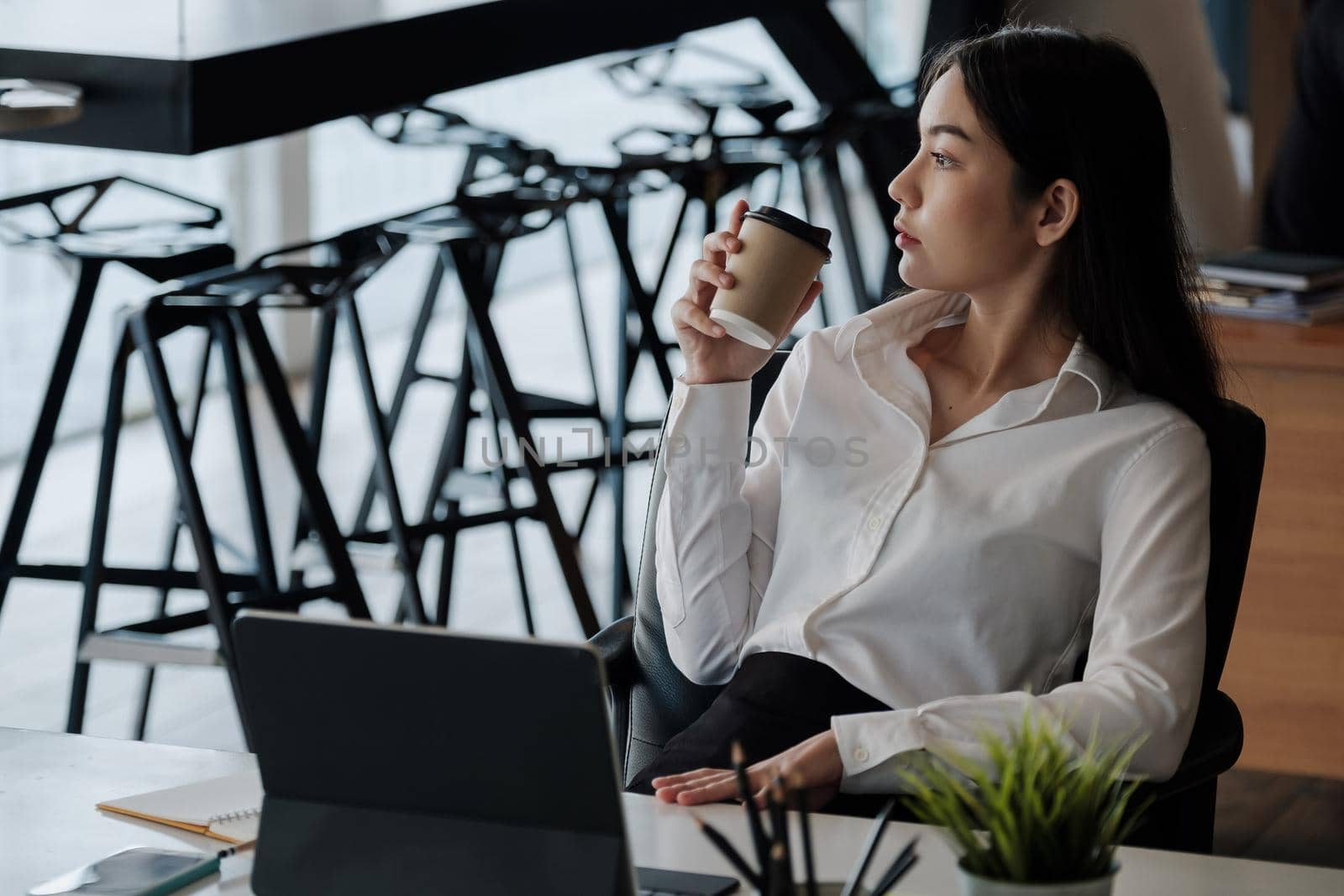 Portrait of beautiful woman or accountant sitting at desk in modern office with interior drinking hot beverage holding cup