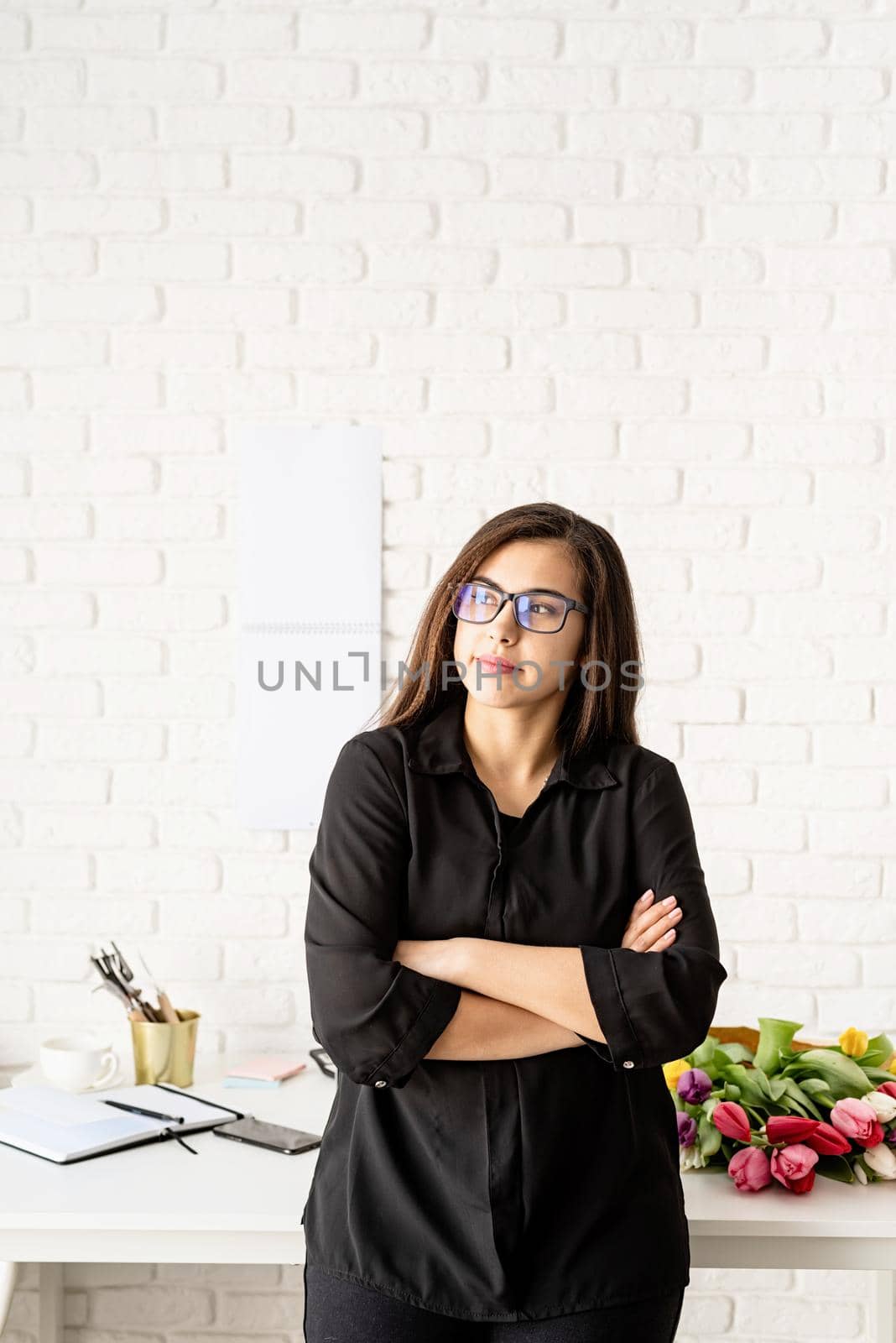 Portrait of young confident brunette business woman working at the office, standing by the desk with arms crossed by Desperada