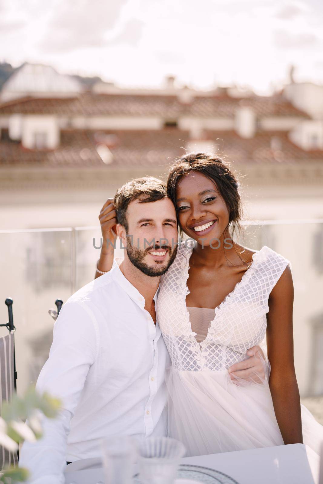 Interracial wedding couple. Destination fine-art wedding in Florence, Italy. African-American bride and Caucasian groom are sitting at rooftop wedding dinner table overlooking the city.