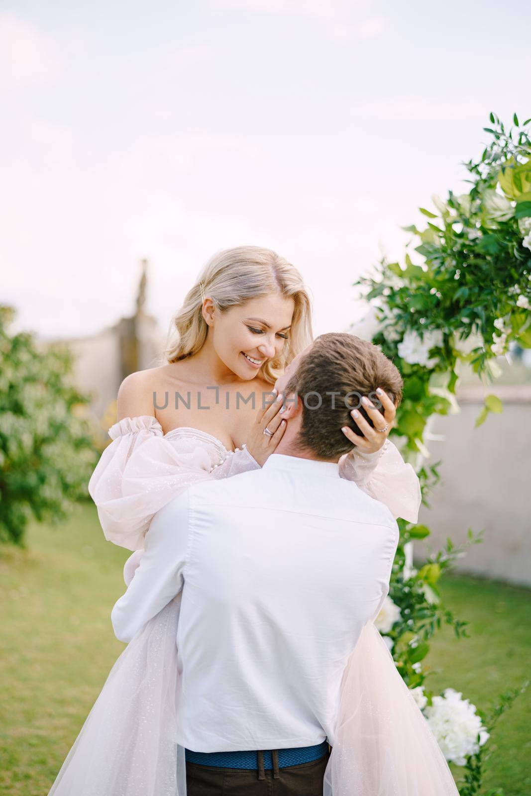 Wedding at an old winery villa in Tuscany, Italy. The groom circles the bride in his arms. Round wedding arch decorated with white flowers and greenery in front of an ancient Italian architecture. by Nadtochiy