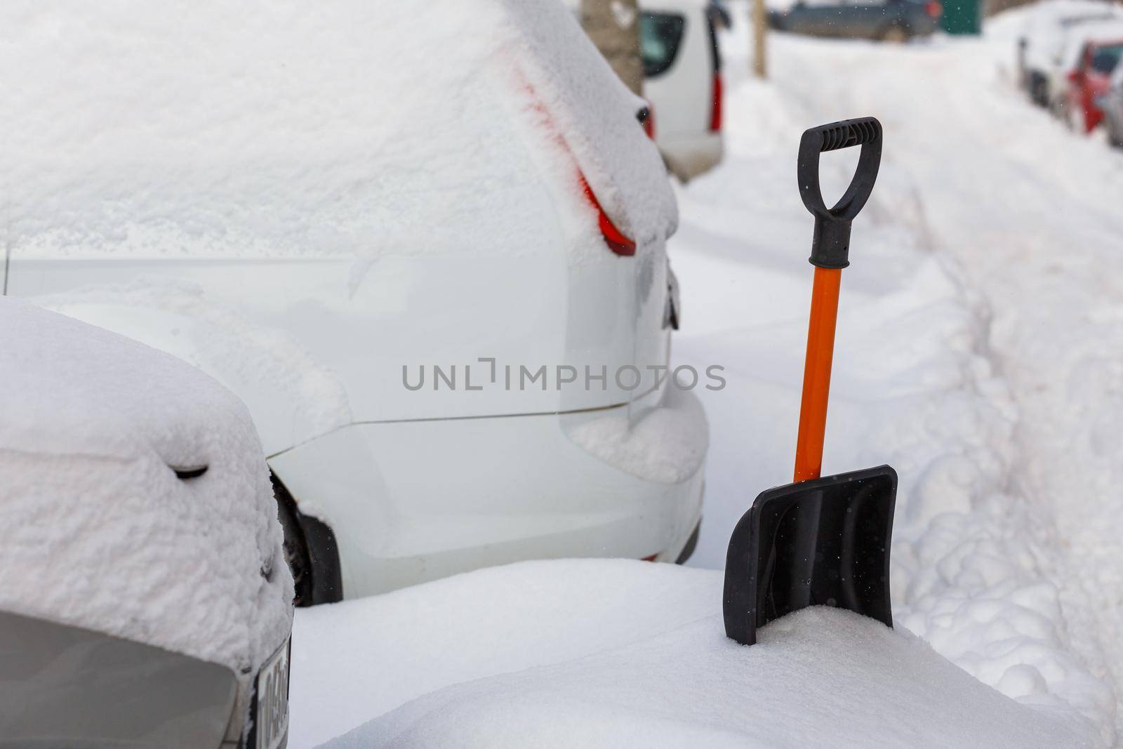 small plastic snow shovel near snow-covered cars at sunny winter morning while people cleaning their cars out of snow
