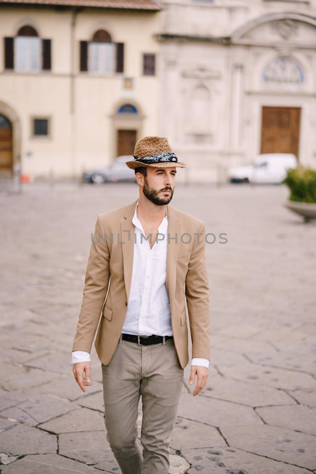 A young guy with a straw hat, a white unbuttoned shirt and a sand jacket, a small beard walks around the city of Florence. by Nadtochiy