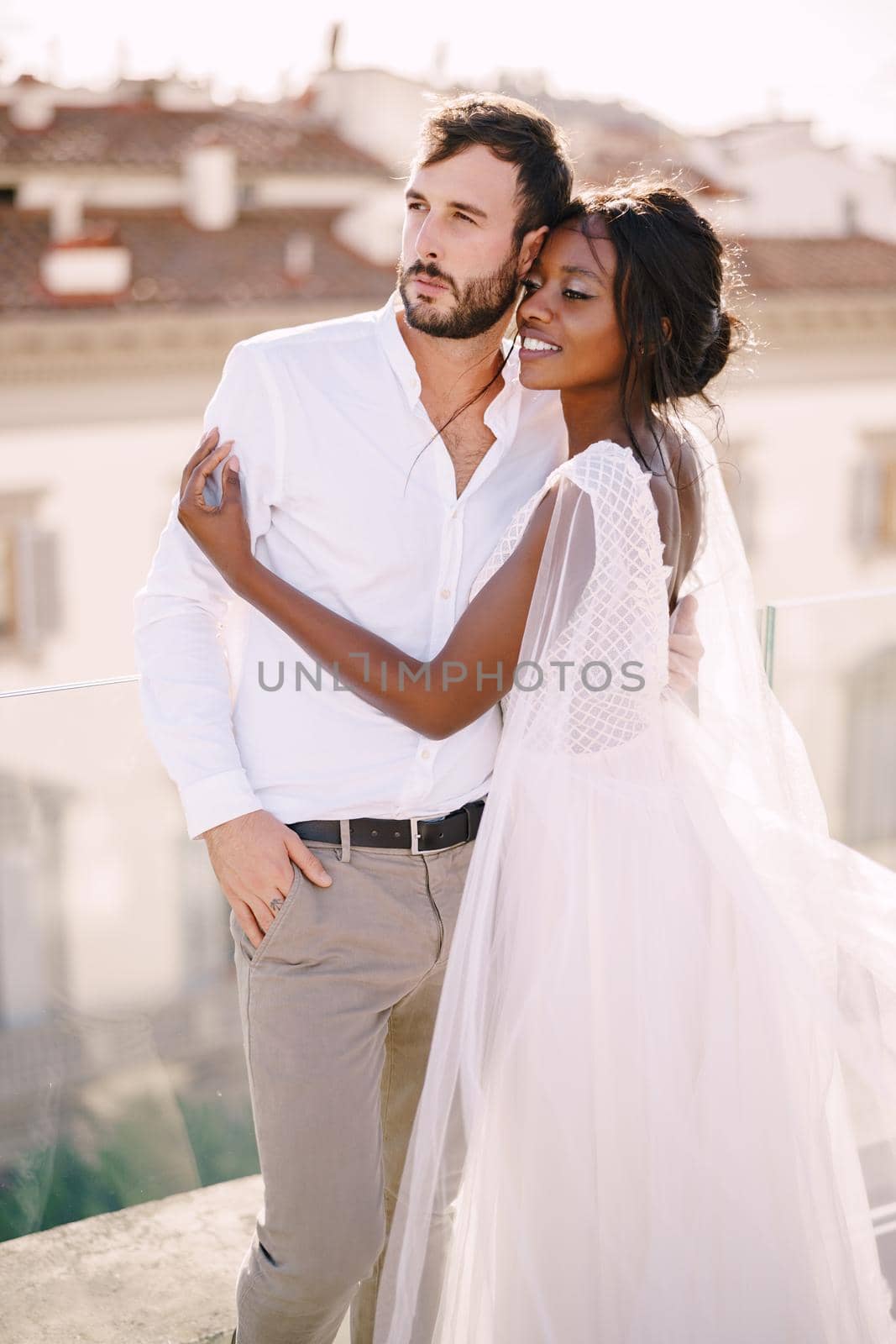 Interracial wedding couple. Destination fine-art wedding in Florence, Italy. African-American bride hugs Caucasian groom on the roof with cityscape view. by Nadtochiy