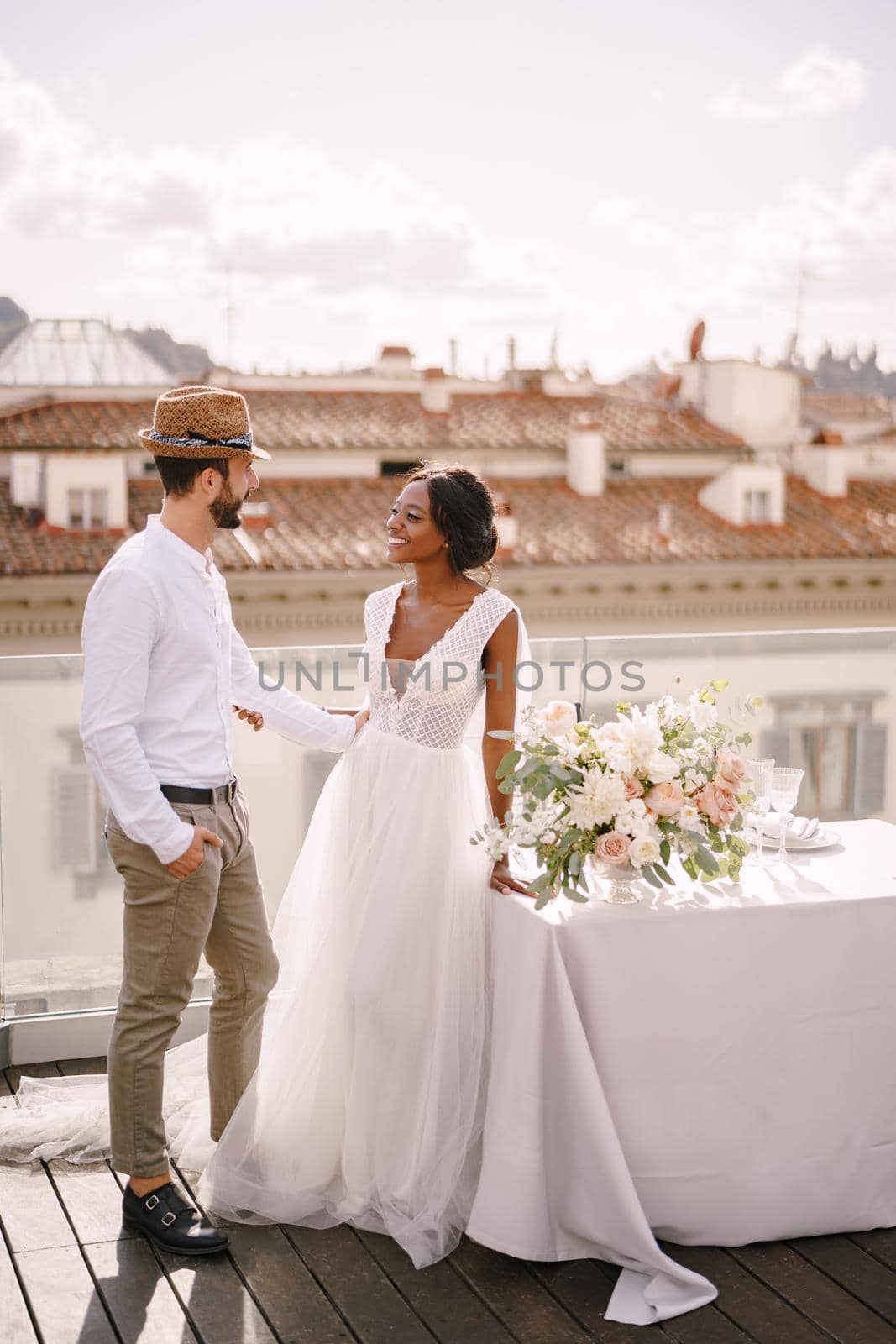 Interracial wedding couple. Destination fine-art wedding in Florence, Italy. African-American bride and Caucasian groom with a beard and straw hat, near the table for a wedding dinner. by Nadtochiy