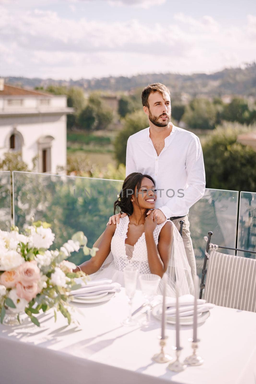 Mixed-race wedding couple. Destination fine-art wedding in Florence, Italy. African-American bride sits at wedding table, Caucasian groom hugs her shoulders.