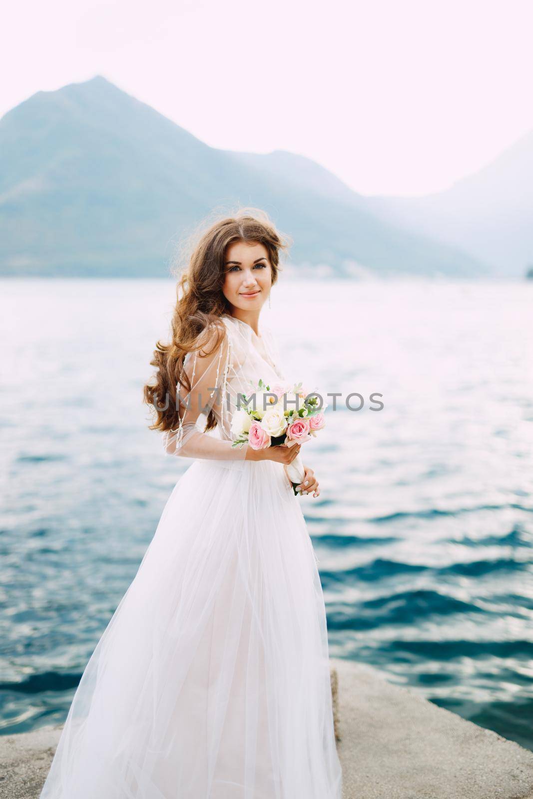 The bride holds a bouquet of roses in her hands and stands on the pier in the Bay of Kotor by Nadtochiy