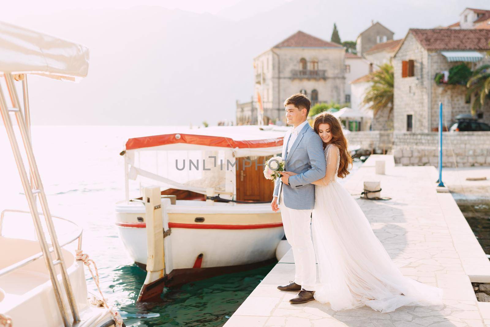 The bride hugs the groom from behind on the pier near the old town of Perast, next to them is a tourist boat . High quality photo