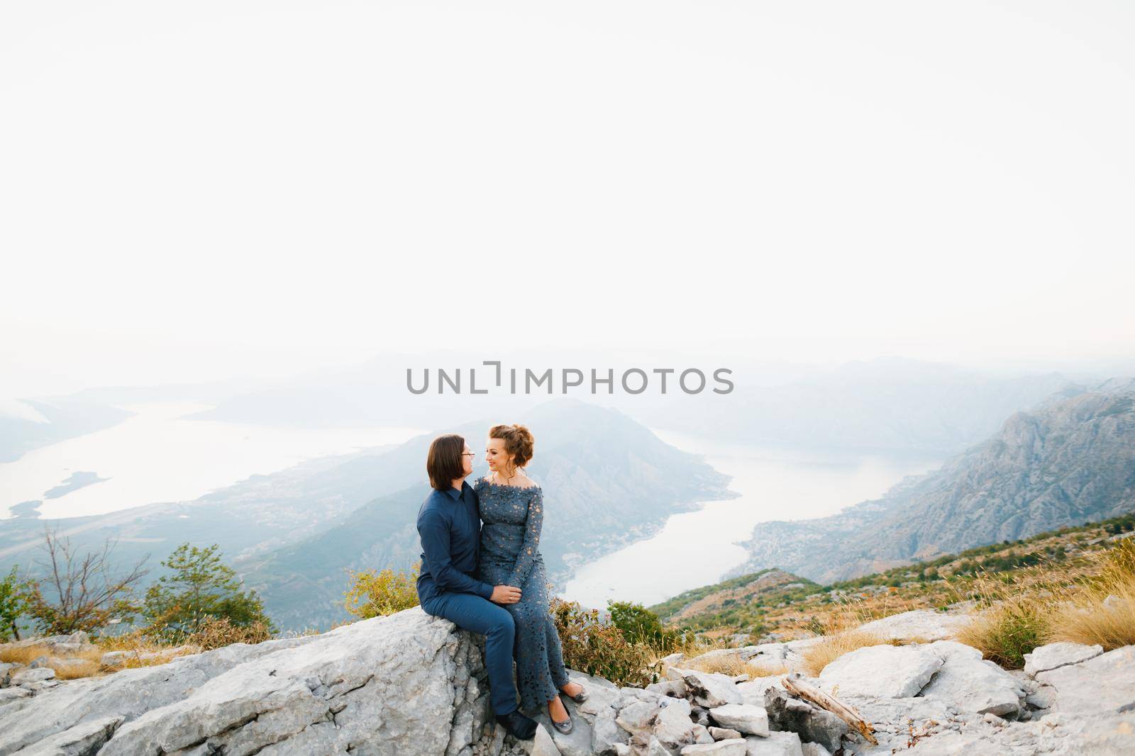 The bride and groom sitting embracing on the Lovcen mountain behind them opens a view of the Bay of Kotor by Nadtochiy