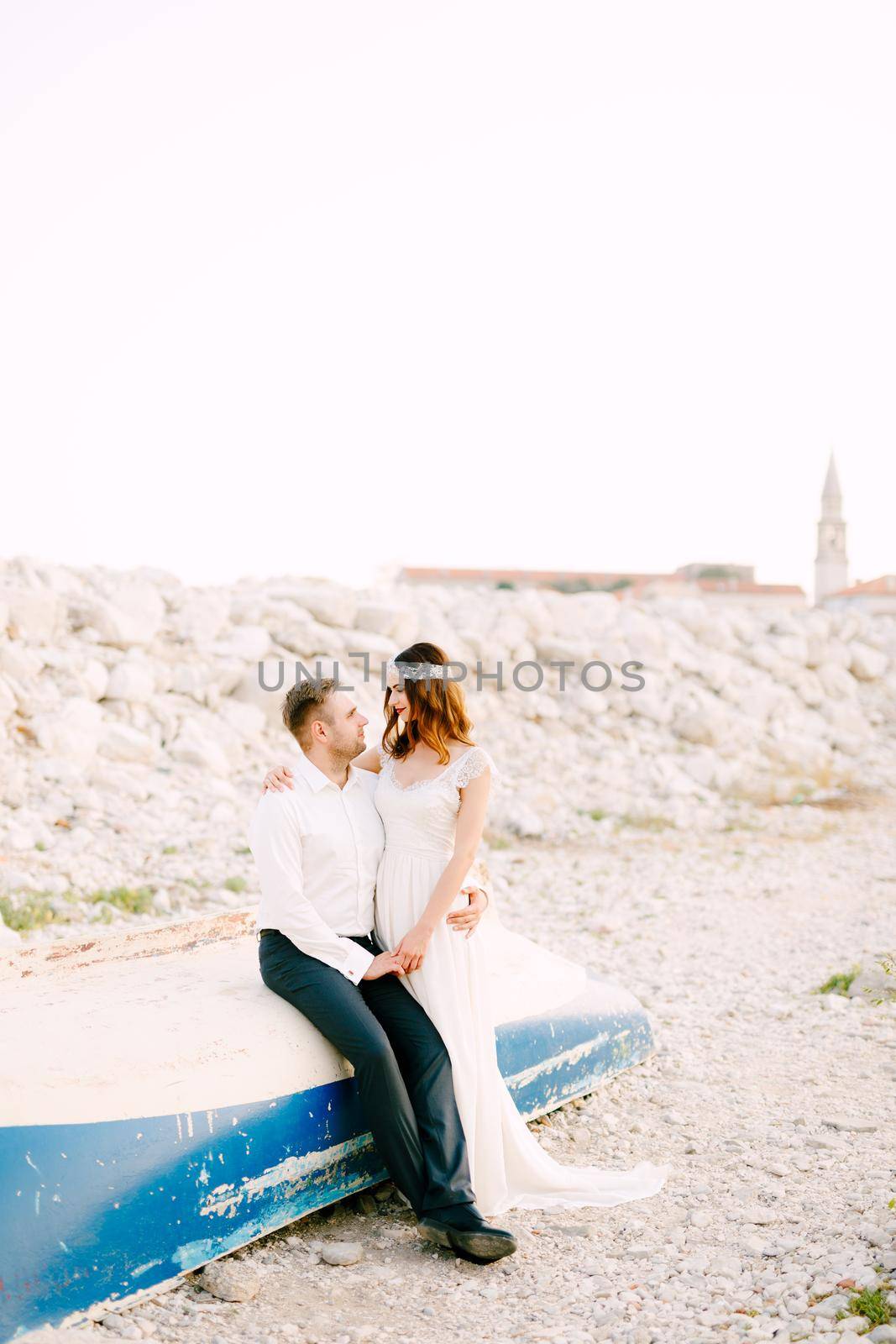 Bride and groom sit embracing on an inverted boat on the beach near old town of Budva . High quality photo