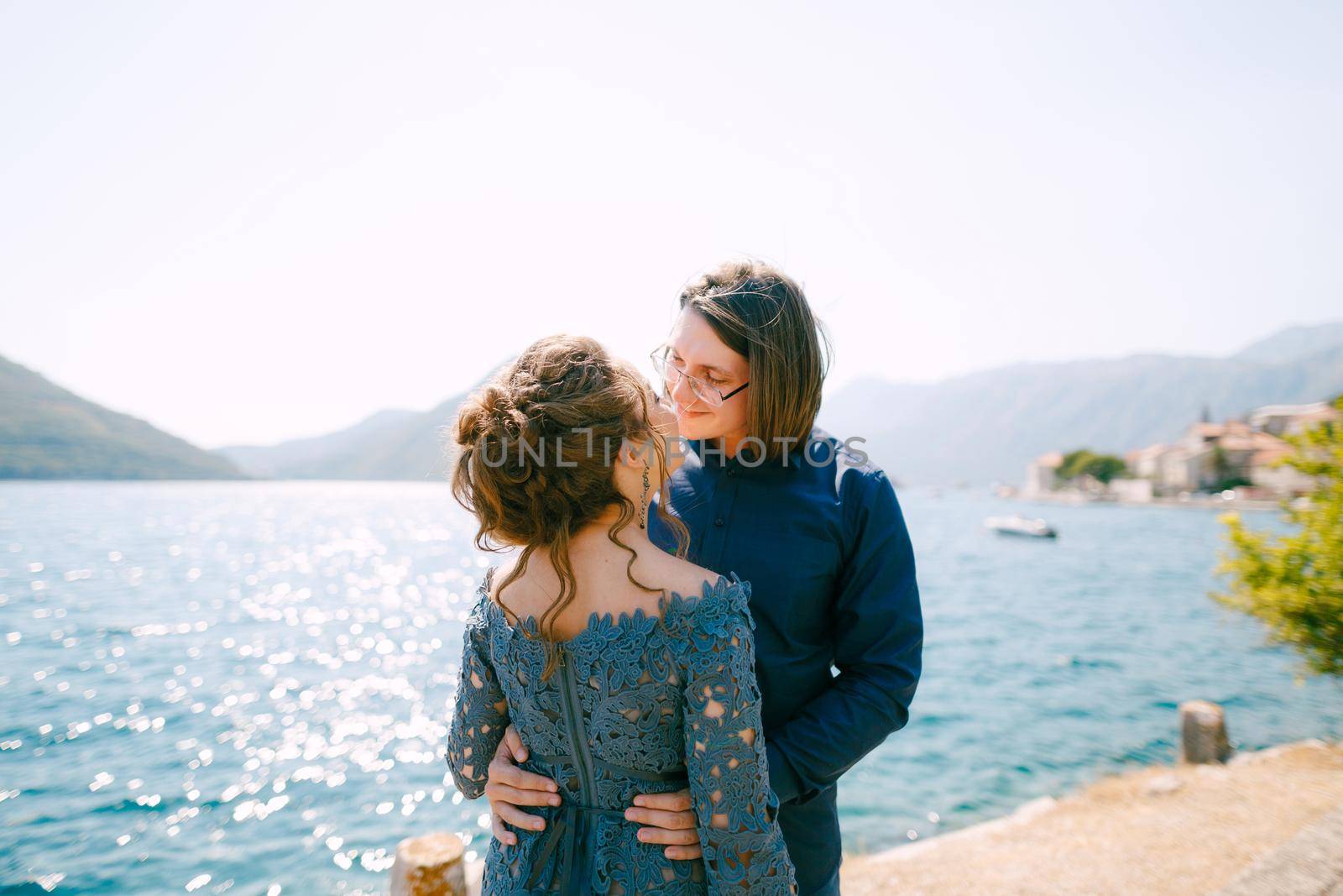 The bride in stylish grey dress and groom hugging on the pier near the old town of Perast by Nadtochiy