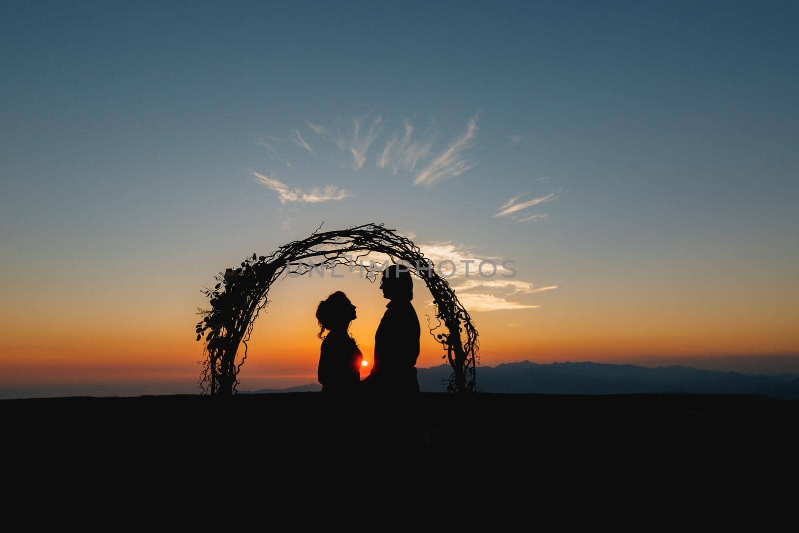 Silhouettes of bride and groom holding hands near the wedding arch at sunset at the wedding ceremony by Nadtochiy