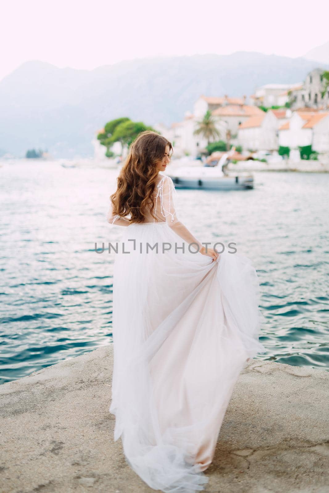 The bride stands on the pier near the old town of Perast and holds the edge of her skirt. High quality photo