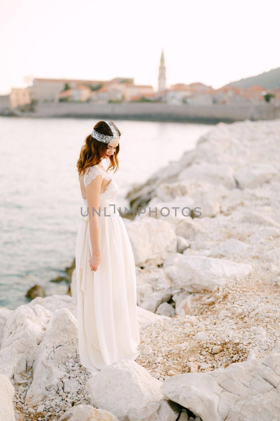 A sophisticated bride stands on a picturesque rocky shore near the old town of Budva by Nadtochiy