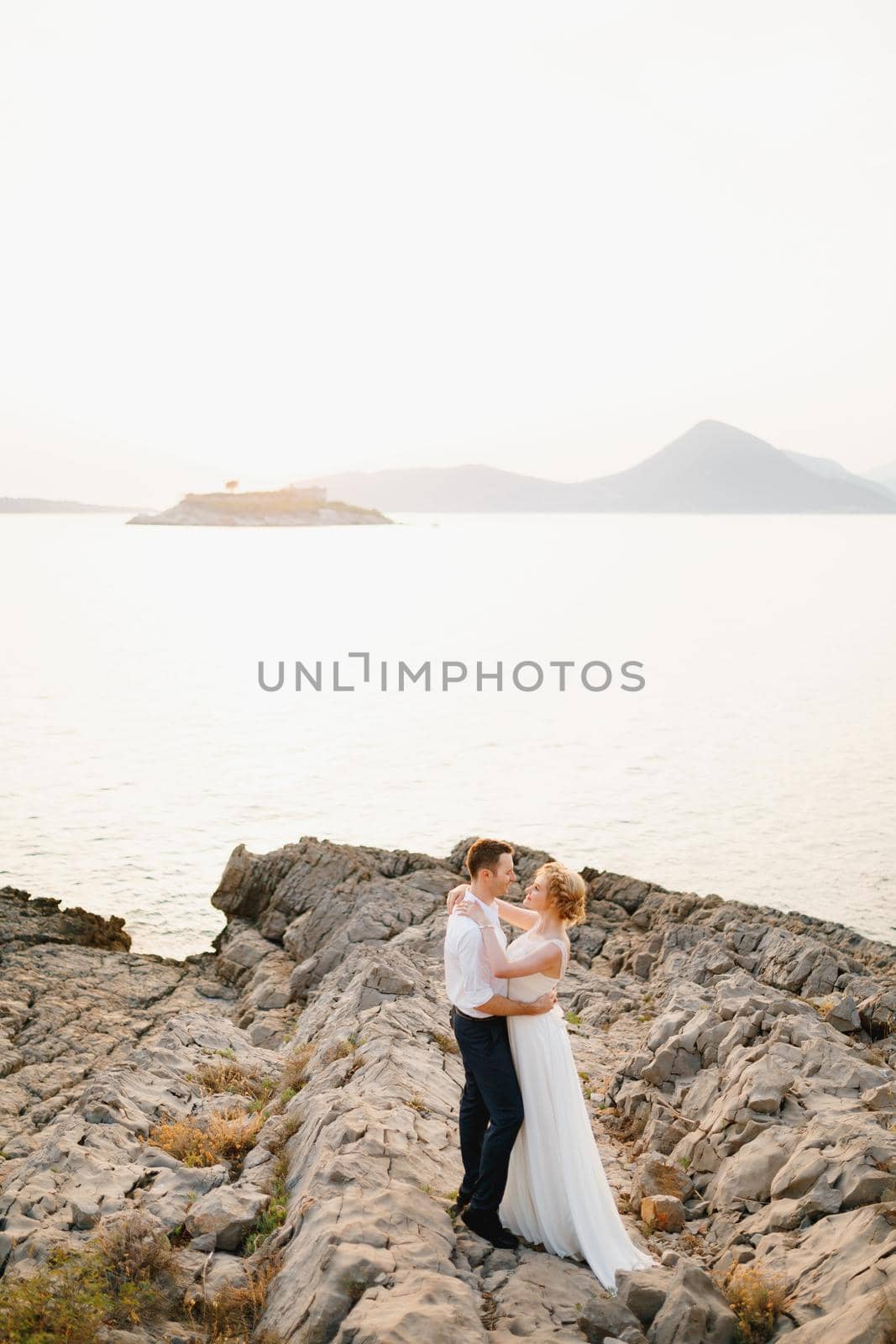 The bride and groom hug on the rocks by the sea against the backdrop of the mountains and the island of Mamula by Nadtochiy