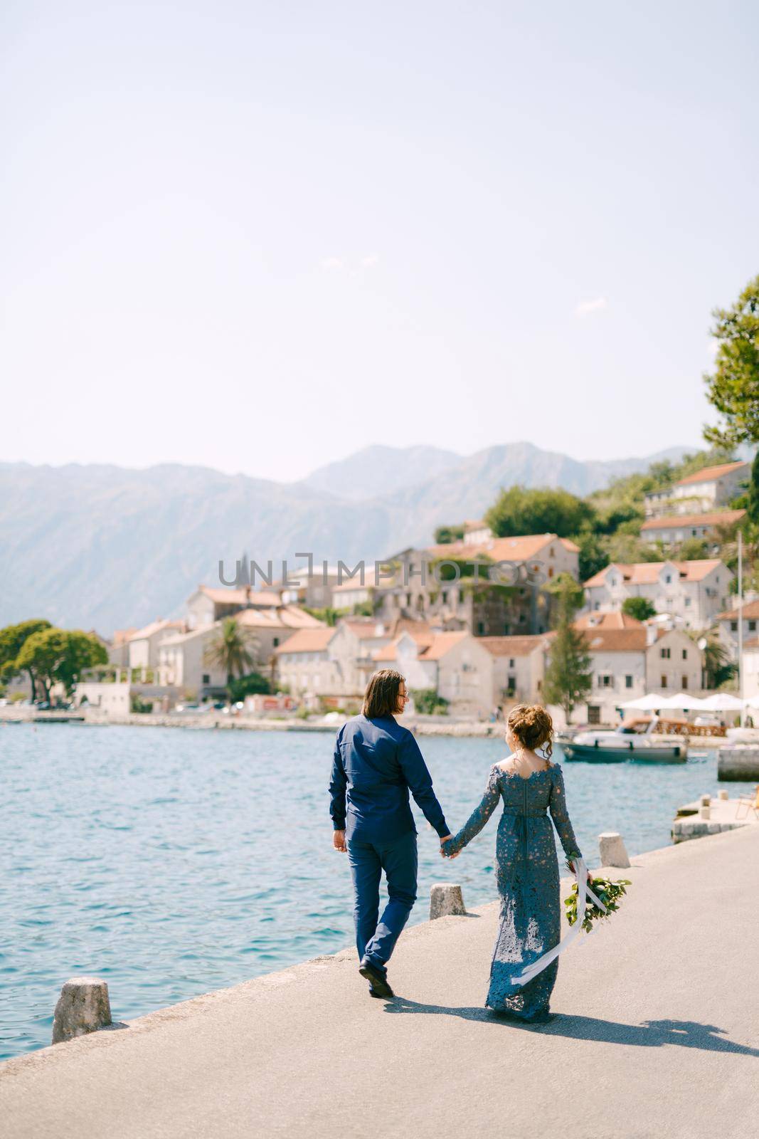 The bride in a stylish blue dress and groom walk along the pier holding hands near the old town of Perast, back view . High quality photo