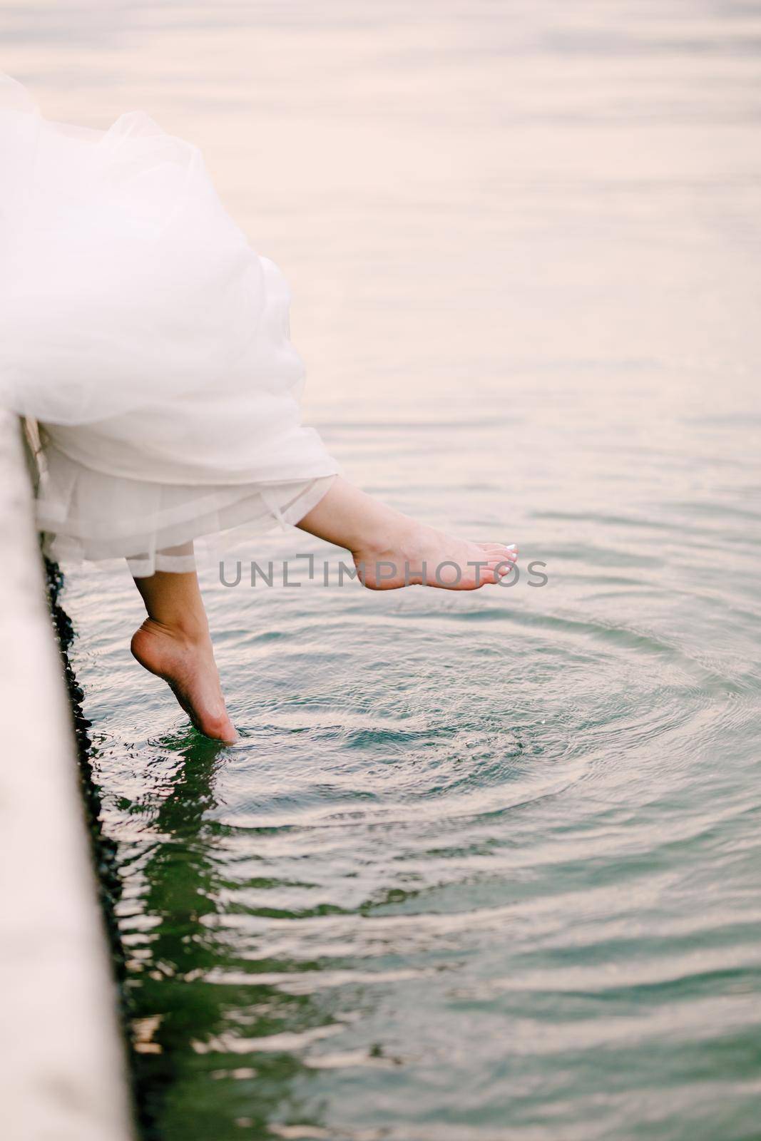 The bride sits on the pier and dangles her bare feet in the water, the legs peek out from under the wedding dress by Nadtochiy