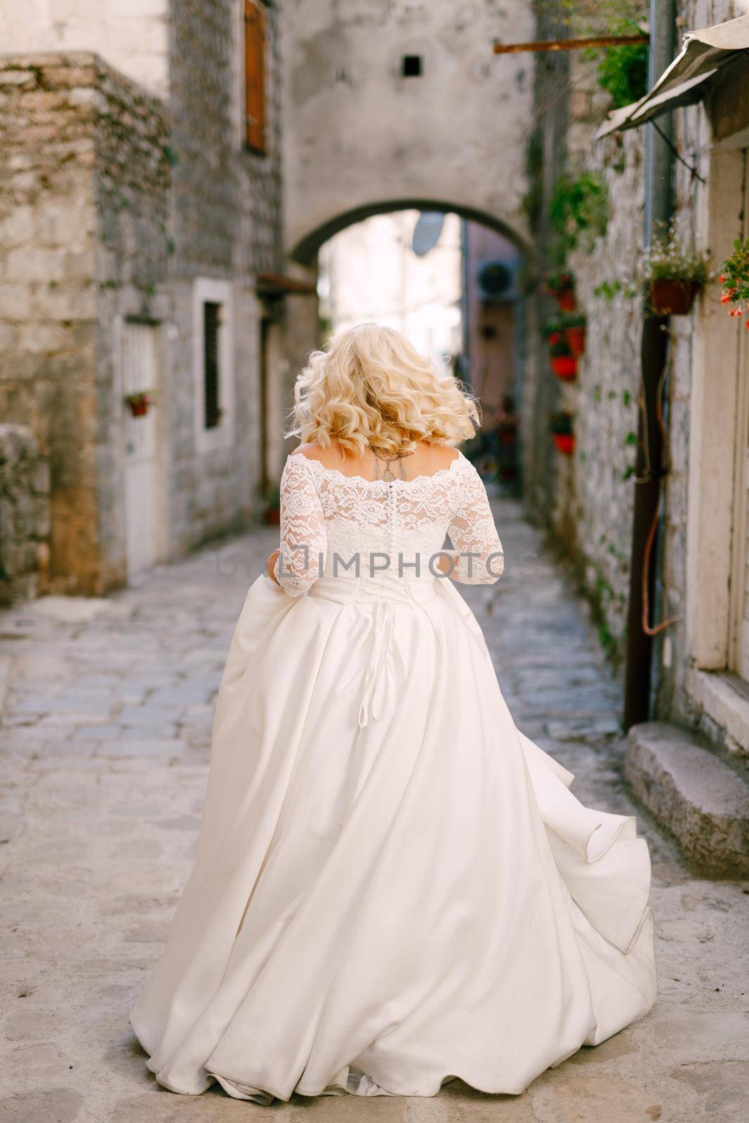 The bride walks along a beautiful narrow street of the old town of Perast with brick houses, back view . High quality photo