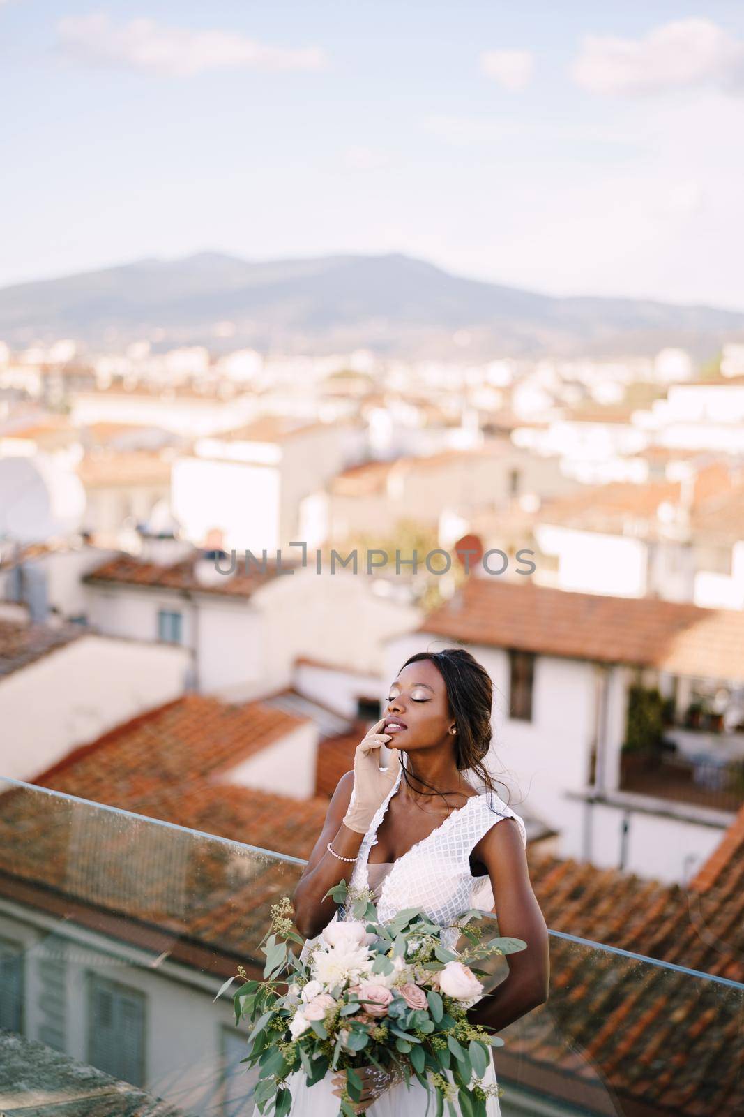 An African-American bride in a white dress, stands on the roof terrace, with a bouquet in her hands and in short gloves. Fine-art wedding in Florence, Italy