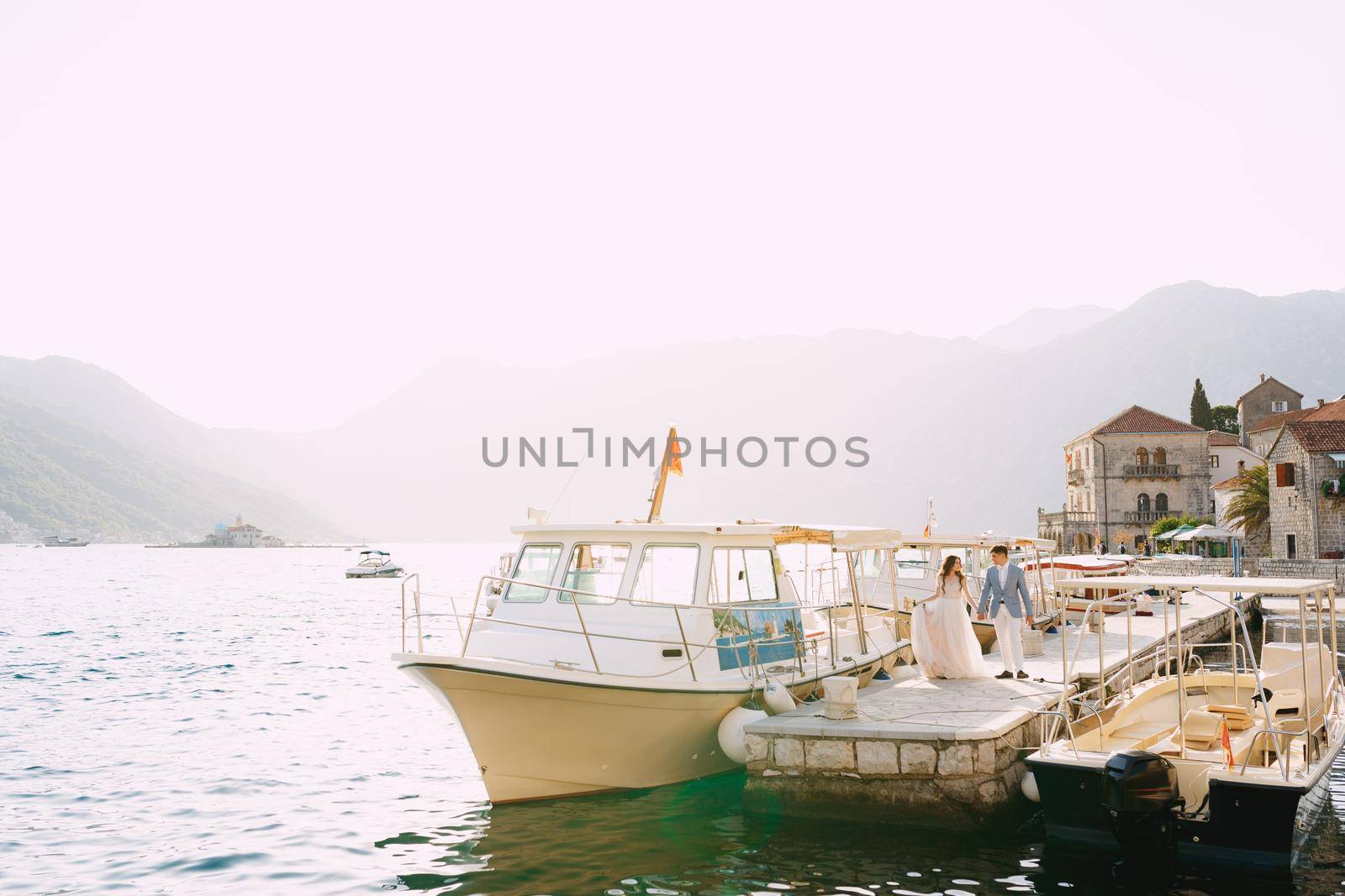 The bride and groom walk holding hands along the pier near the old town of Perast, next to them are tourist boats . High quality photo
