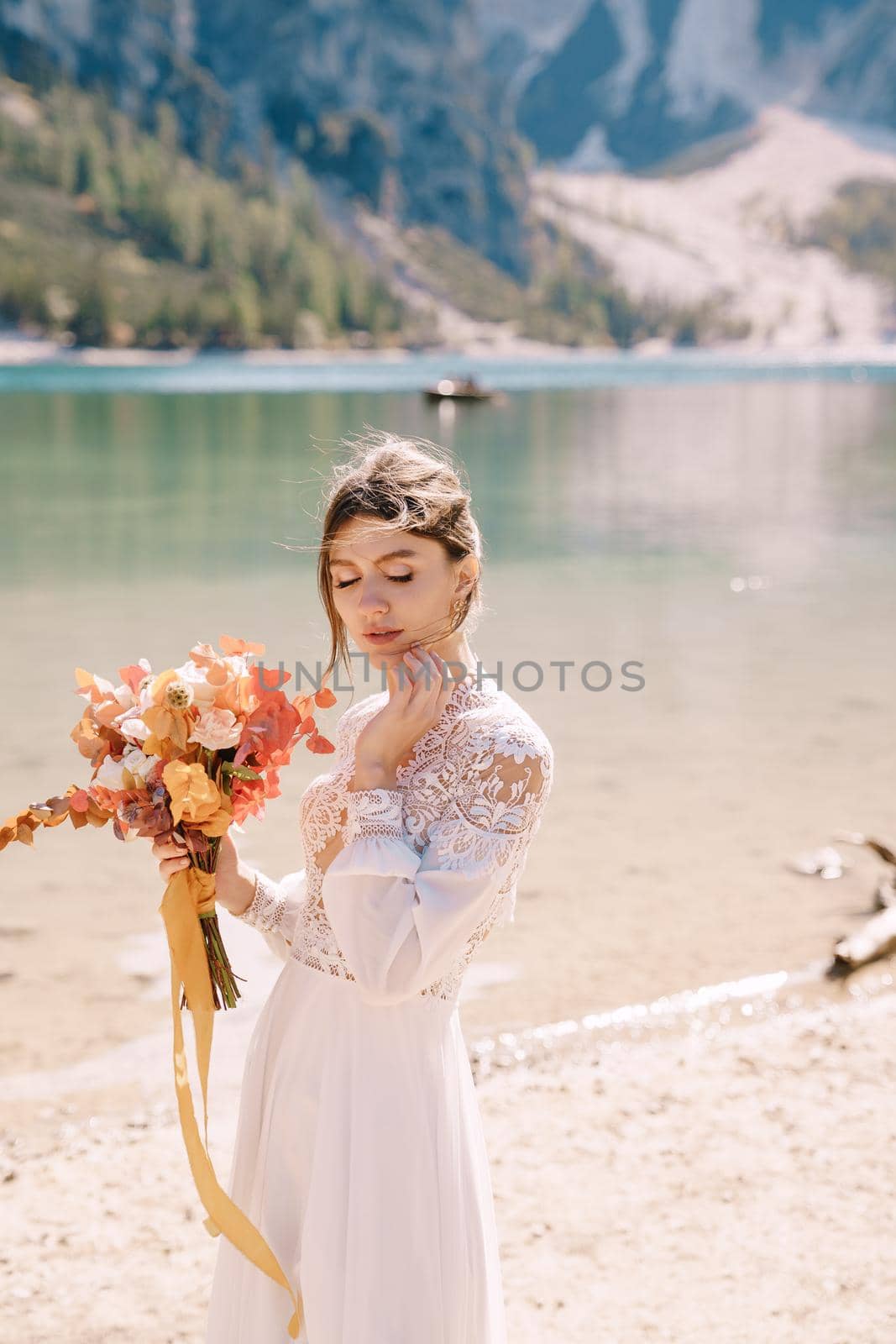 Beautiful bride in a white dress with sleeves and lace, with a yellow autumn bouquet of dried flowers and peony roses, at Lago di Braies in Italy. Destination wedding in Europe, at Braies lake.