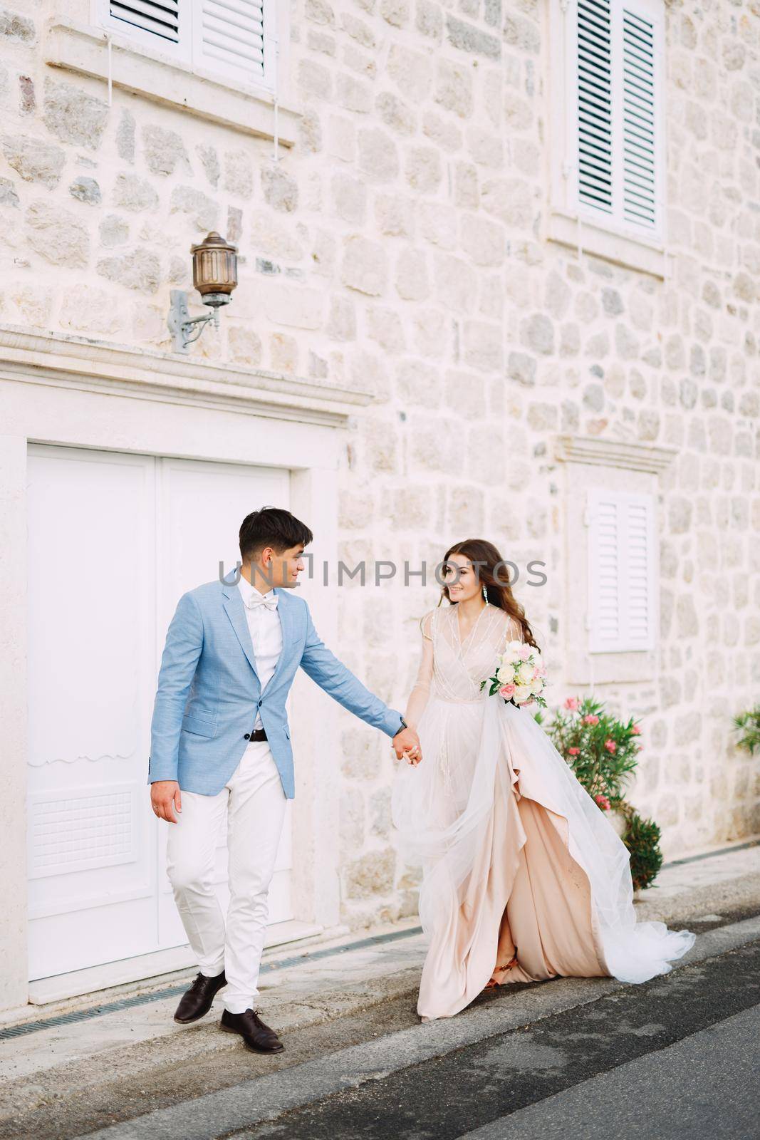 The bride and groom stand holding hands in front of a beautiful white house in the old town of Perast by Nadtochiy