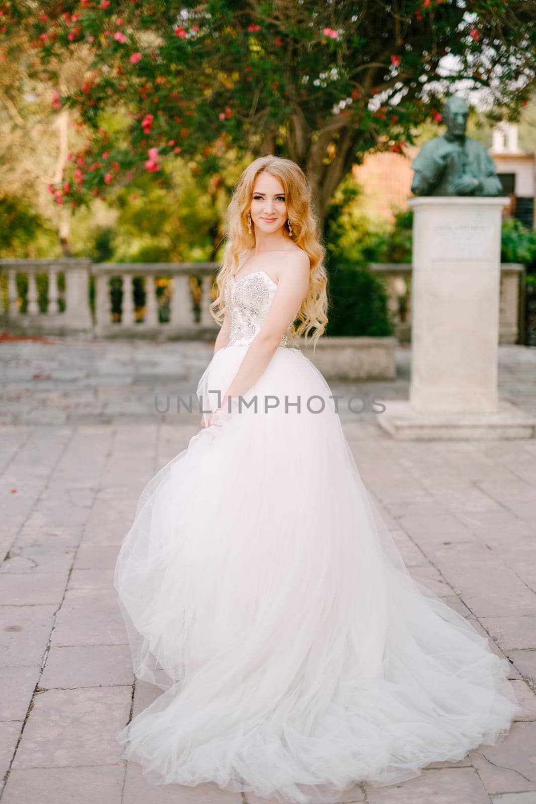 A blonde-haired bride in a lush white dress stands on the site near the ancient church in Prcanj by Nadtochiy