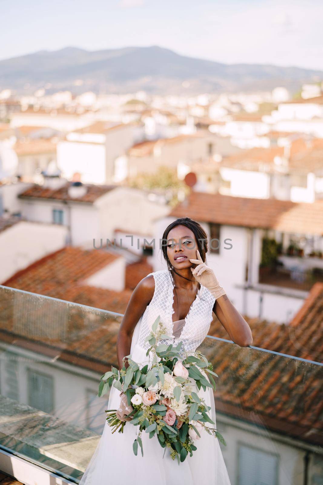 An African-American bride in a white dress, stands on the roof terrace, with a bouquet in her hands and in short gloves. Fine-art wedding in Florence, Italy
