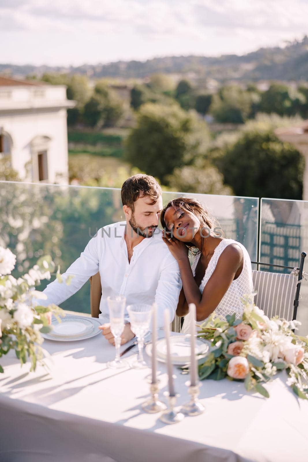 Destination fine-art wedding in Florence, Italy. African-American bride and Caucasian groom are sitting at the rooftop wedding dinner table overlooking the city. Multiracial wedding couple by Nadtochiy