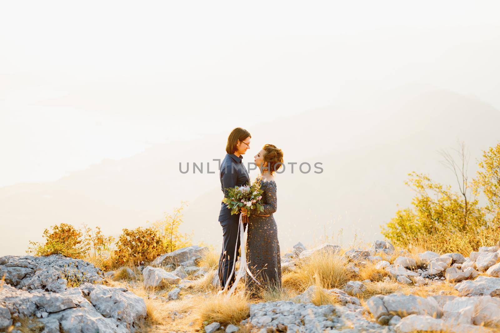 The bride and groom embracing and looking at each other on the Lovcen mountain behind them opens a view of the Bay of Kotor by Nadtochiy