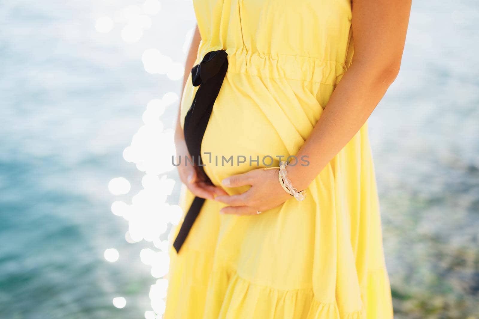 A pregnant woman in a cute bright yellow dress stands by the sea and holds her belly with her hands, close-up by Nadtochiy