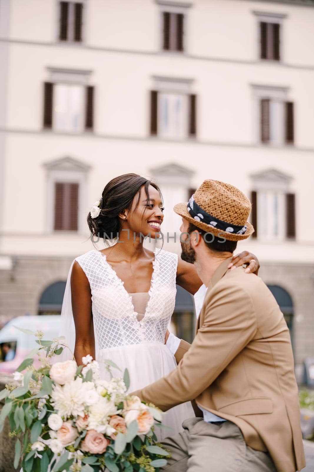 Multiracial wedding couple. Wedding in Florence, Italy. African-American bride in a white dress with a long veil and a bouquet, and Caucasian groom in a sandy jacket and straw hat. by Nadtochiy