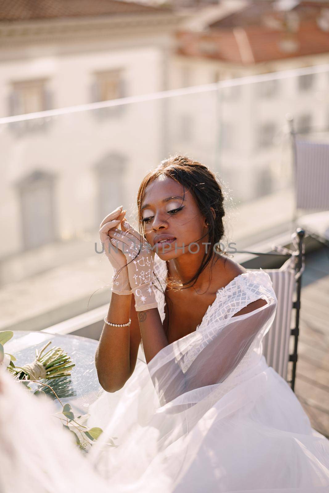 Destination fine-art wedding in Florence, Italy. African-American bride sits at the table, touches his face with her hands in gloves, a bouquet lies on the table.