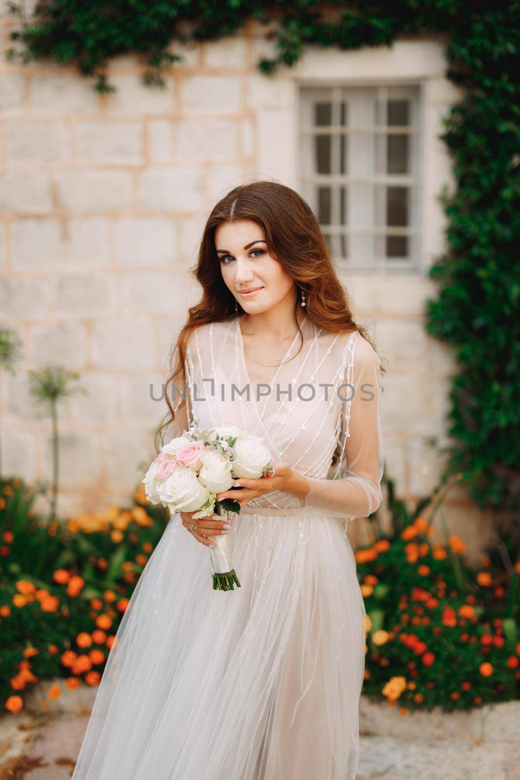 A bride with a bouquet in her hands stands at the wall of a house with a green liana and orange flowers in Perast . High quality photo