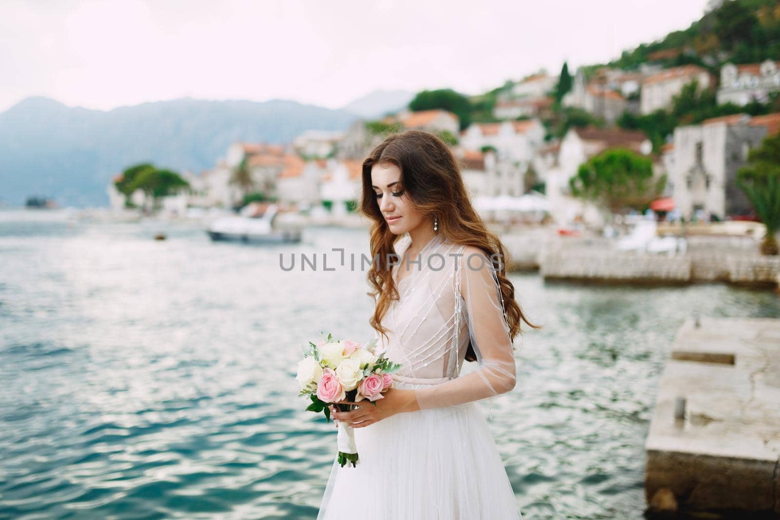 The bride holds a bouquet of roses in her hands and stands on the pier near the old town of Perast . High quality photo