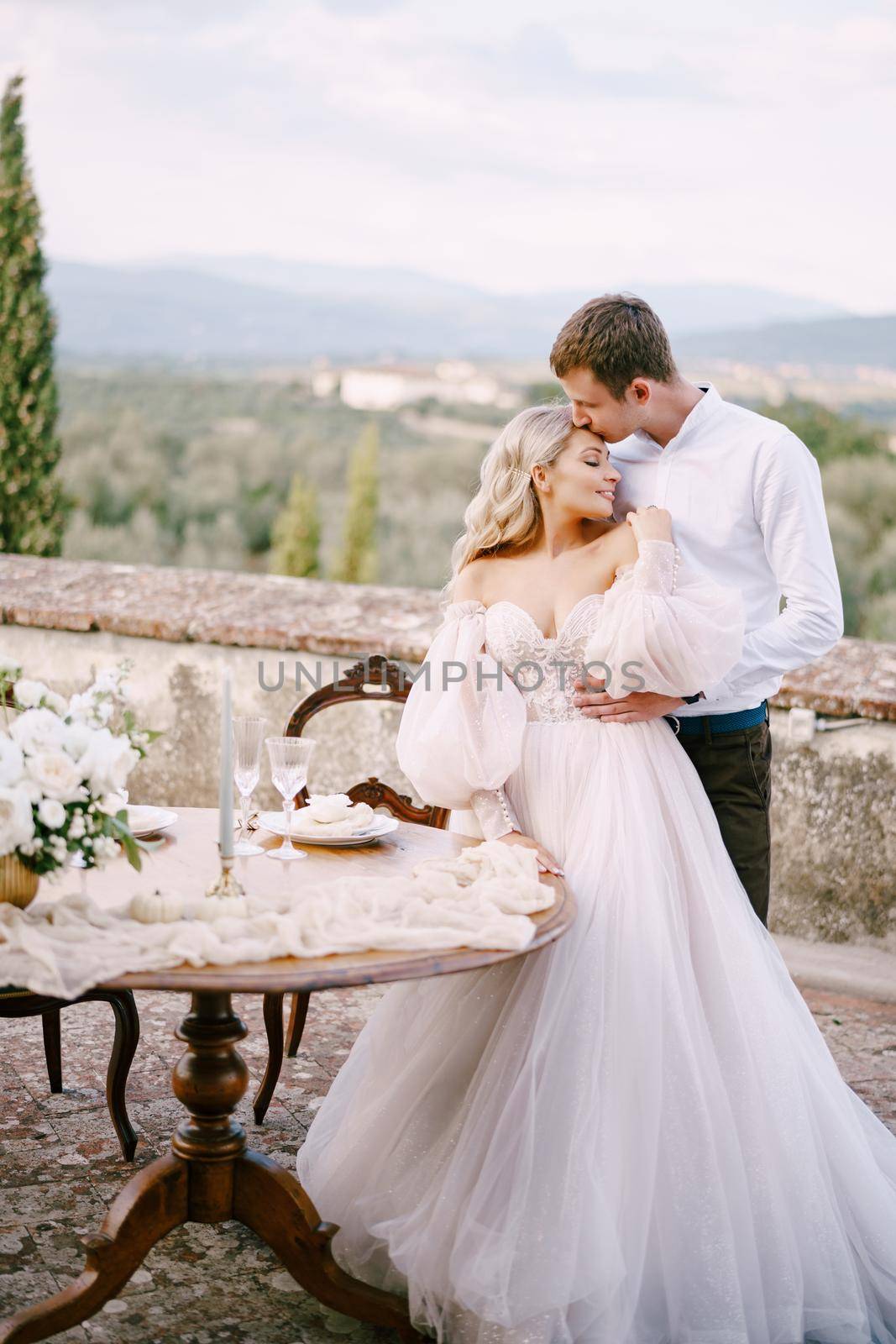 Wedding at an old winery villa in Tuscany, Italy. The wedding couple stands near the table for the wedding dinner, the groom hugs the bride at the waist.