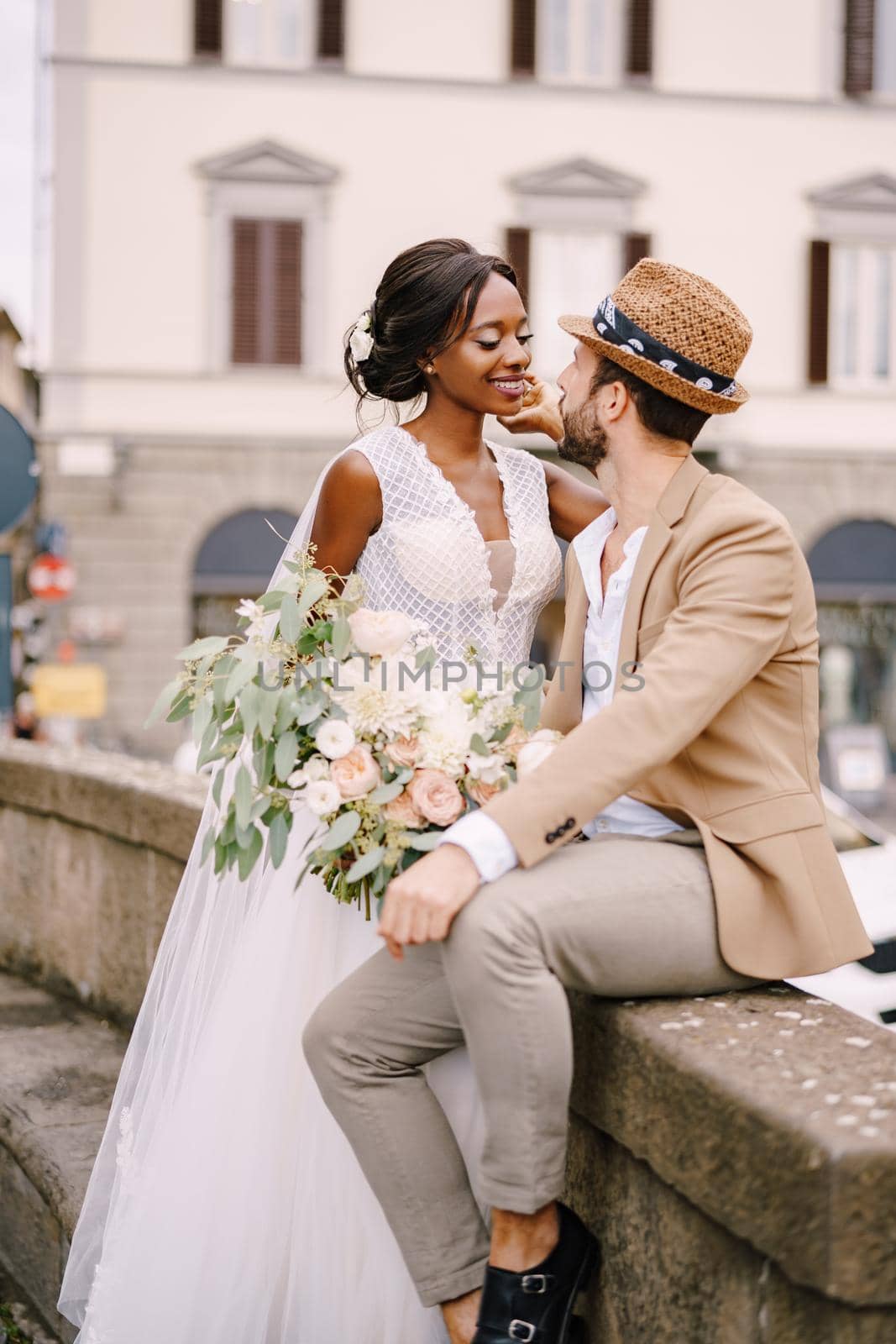 Wedding in Florence, Italy. Interracial wedding couple. African-American bride in a white dress with a long veil and a bouquet, and Caucasian groom in a sandy jacket and straw hat. by Nadtochiy