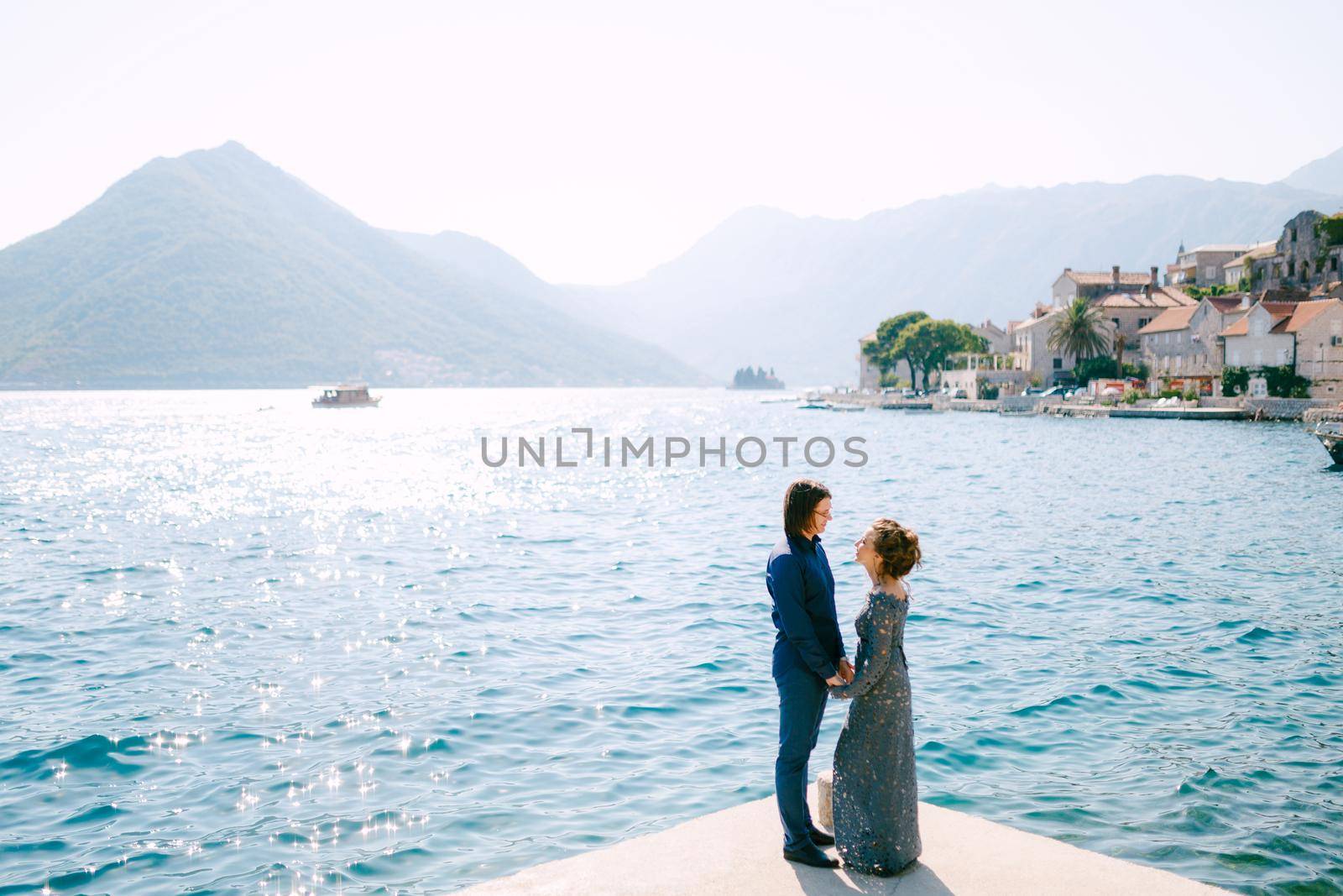 The bride in a stylish grey wedding dress and the groom stand holding hands on the seashore near the old town of Perast. High quality photo