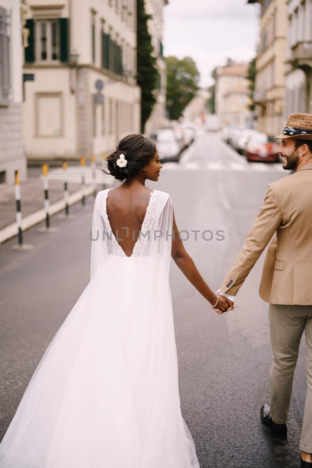 Multiracial wedding couple. Wedding in Florence, Italy. African-American bride in a white dress and Caucasian groom in a hat are walking along the road among cars. by Nadtochiy