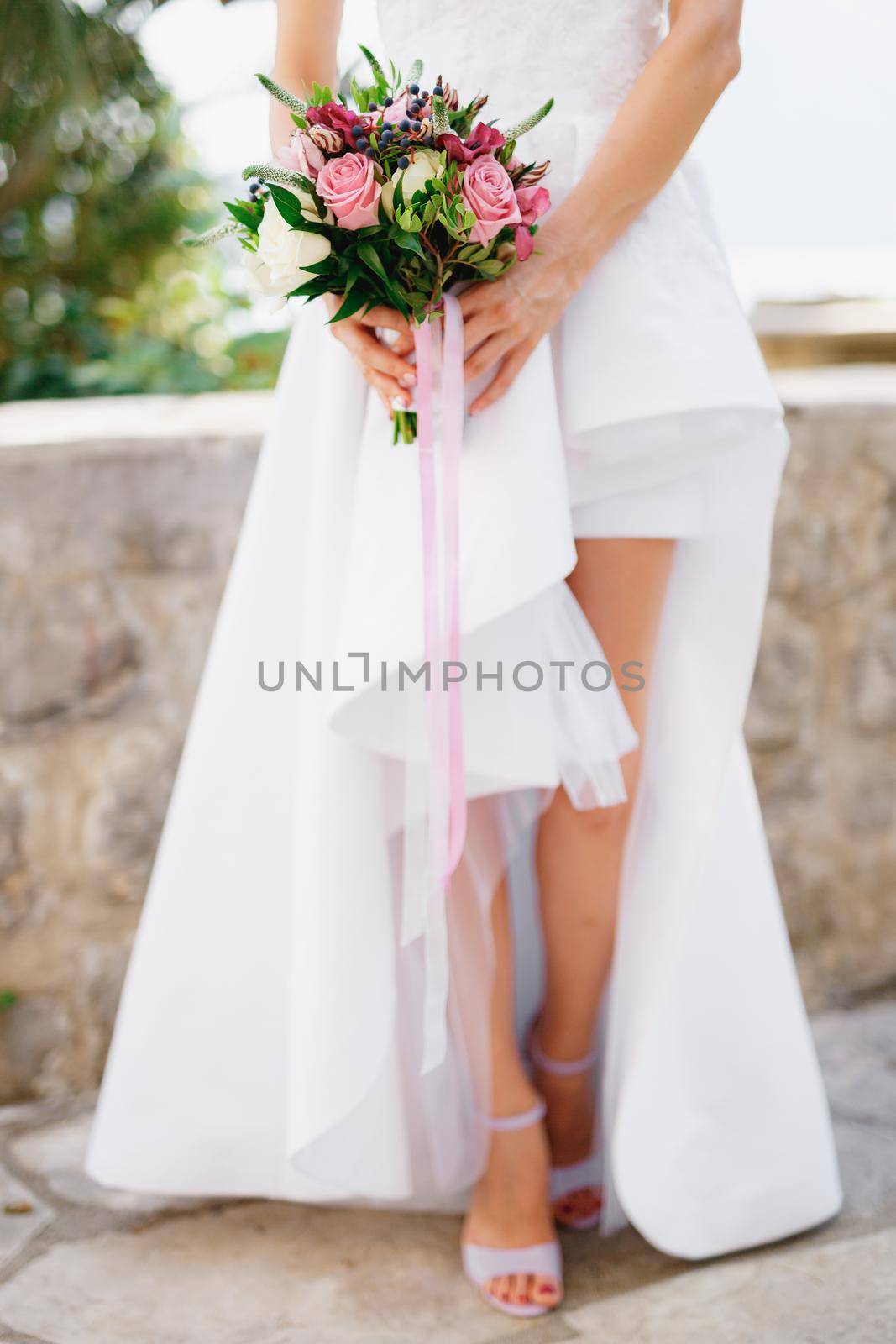 Bride in a stylish dress with open legs holding a wedding bouquet with roses, veronica, viburnum and boxwood in her hands, close-up by Nadtochiy