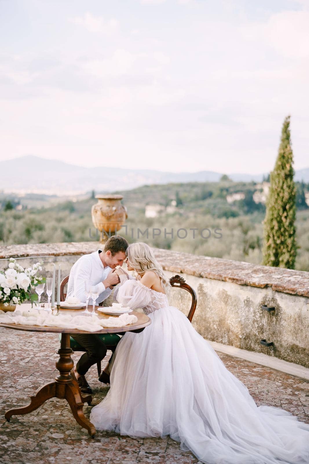 The wedding couple sits at the dinner table on the roof of the old villa, the groom holds the bride's hands. Wedding at an old winery villa in Tuscany, Italy