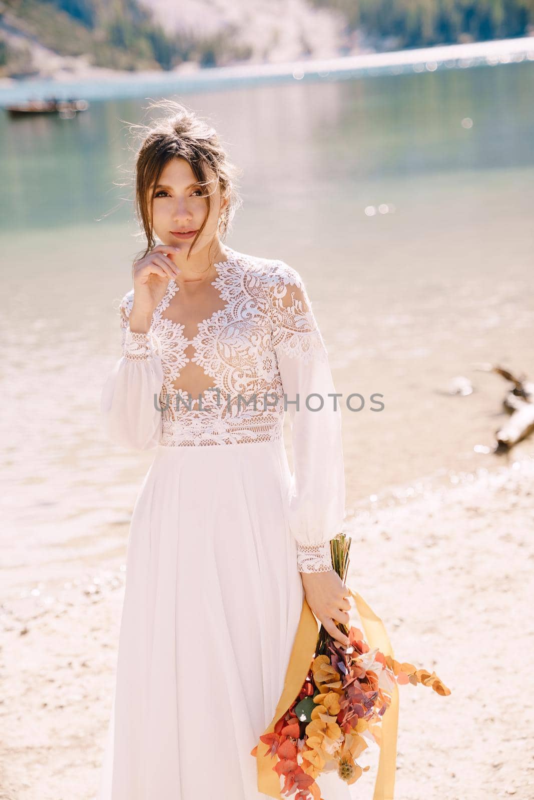 Beautiful bride in a white dress with sleeves and lace, with a yellow autumn bouquet of dried flowers and peony roses, at Lago di Braies in Italy. Destination wedding in Europe, at Braies lake.